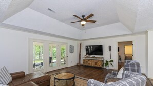 A spacious living room with a brown leather sofa, grey plaid armchairs, a flat-screen TV, wooden cabinets, and a ceiling fan.