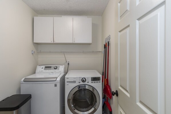 A laundry room with a front-loading washer and dryer, a cabinet above, and a broom leaning against the wall.