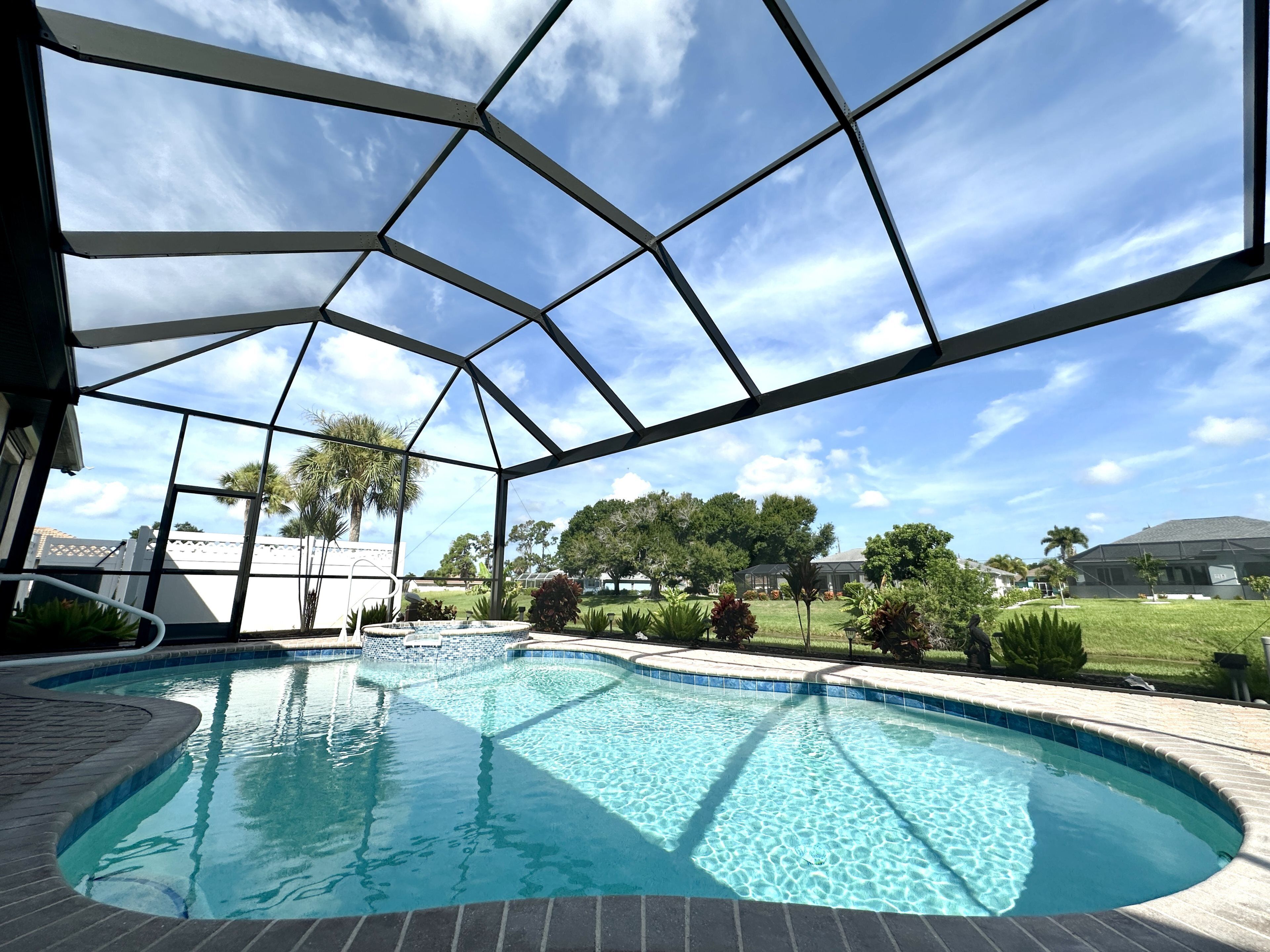 A swimming pool with a clear blue water, surrounded by a covered area with a metal frame, and a view of a residential neighborhood with houses and trees.