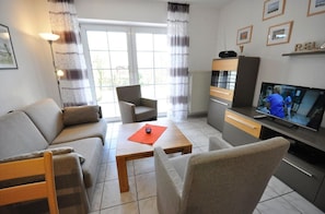 Una sala de estar con un sofá, sillones, una mesa de centro, un televisor y una zona de cocina con un armario y una estantería.