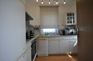 Une cuisine de taille réduite avec des meubles blancs, une cuisinière, un lave-vaisselle, un four à micro-ondes et une fenêtre avec des stores.