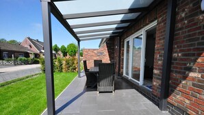 A covered patio with a table and chairs, a brick wall, and a glass door leading to a covered area. - meinStrandhaus - Holiday home "meinStrandhaus" (Neuharlingersiel)