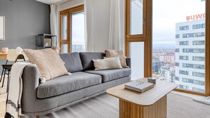 Ein modernes Wohnzimmer mit grauem Sofa, einem Holztisch und Blick auf eine Stadtlandschaft durch große Fenster.