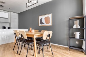 Uma cozinha moderna com armários brancos, uma mesa de jantar de madeira com seis cadeiras e uma parede cinza com um quadro abstrato emoldurado.