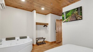 Un dormitorio con una cama grande blanca, techo y piso de madera, bañera blanca, puerta de madera y un cuadro de un puente en un bosque.