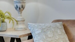 Una sala de estar con un sofá color beige, un cojín estampado, una lámpara blanca con pantalla texturizada y una pequeña planta en maceta sobre una mesita de madera.
