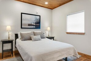 Un dormitorio con cama, dos mesitas de noche, una mesita auxiliar, una ventana con persianas y un cuadro enmarcado en la pared.