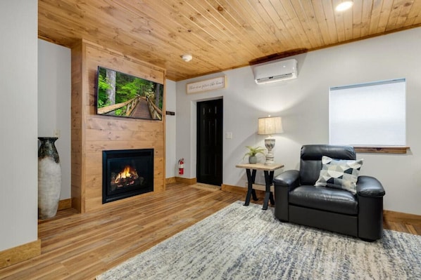 Una sala de estar acogedora con chimenea, un sillón de cuero cómodo, una mesita con lámpara y un cuadro de un sendero en el bosque en la pared.