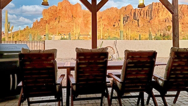 A covered outdoor seating area with wooden chairs and tables, overlooking a desert landscape with cacti and rocky mountains. - Breathtaking Views Spa Dog Friendly w/ Casita (Apache Junction)