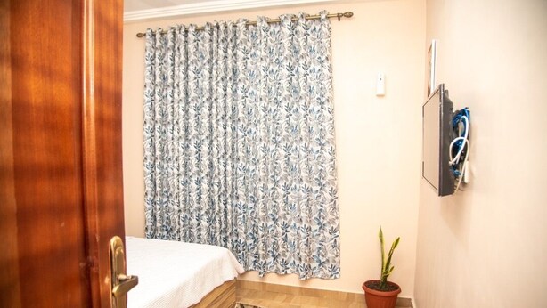 A bedroom with a bed, a patterned curtain, a potted plant, and a wall-mounted TV. - Captivating 1-Bed Apartment in Tema, com 25 (Tema, com 25)