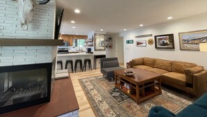 A living room with a fireplace, a sofa, a coffee table, and a rug. There is a kitchen in the background with a bar and stools. - Mid-Century Modern Gem next to Trails, Ski Resorts and Downtown Ogden (Ogden)