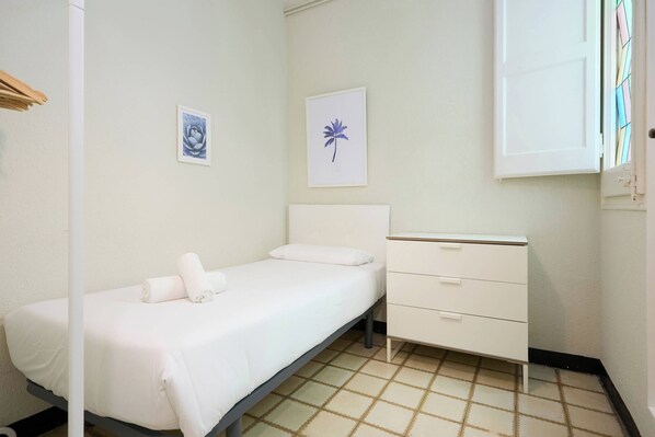 A small, single-bed room with a white bed, a white dresser, and a window with a stained glass design.