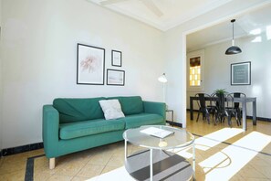 Una sala de estar con un sofá verde, una mesa de centro de vidrio y obras de arte enmarcadas en la pared.