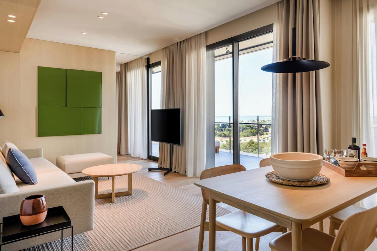 Una habitación de hotel moderna con sofá, una mesa de comedor con un tazón, televisor de pantalla plana y vistas de la ciudad.