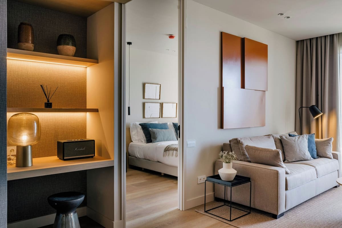Una sala de estar moderna con un sofá, una mesita de centro y una lámpara. Se ve una cama con almohadas al fondo.