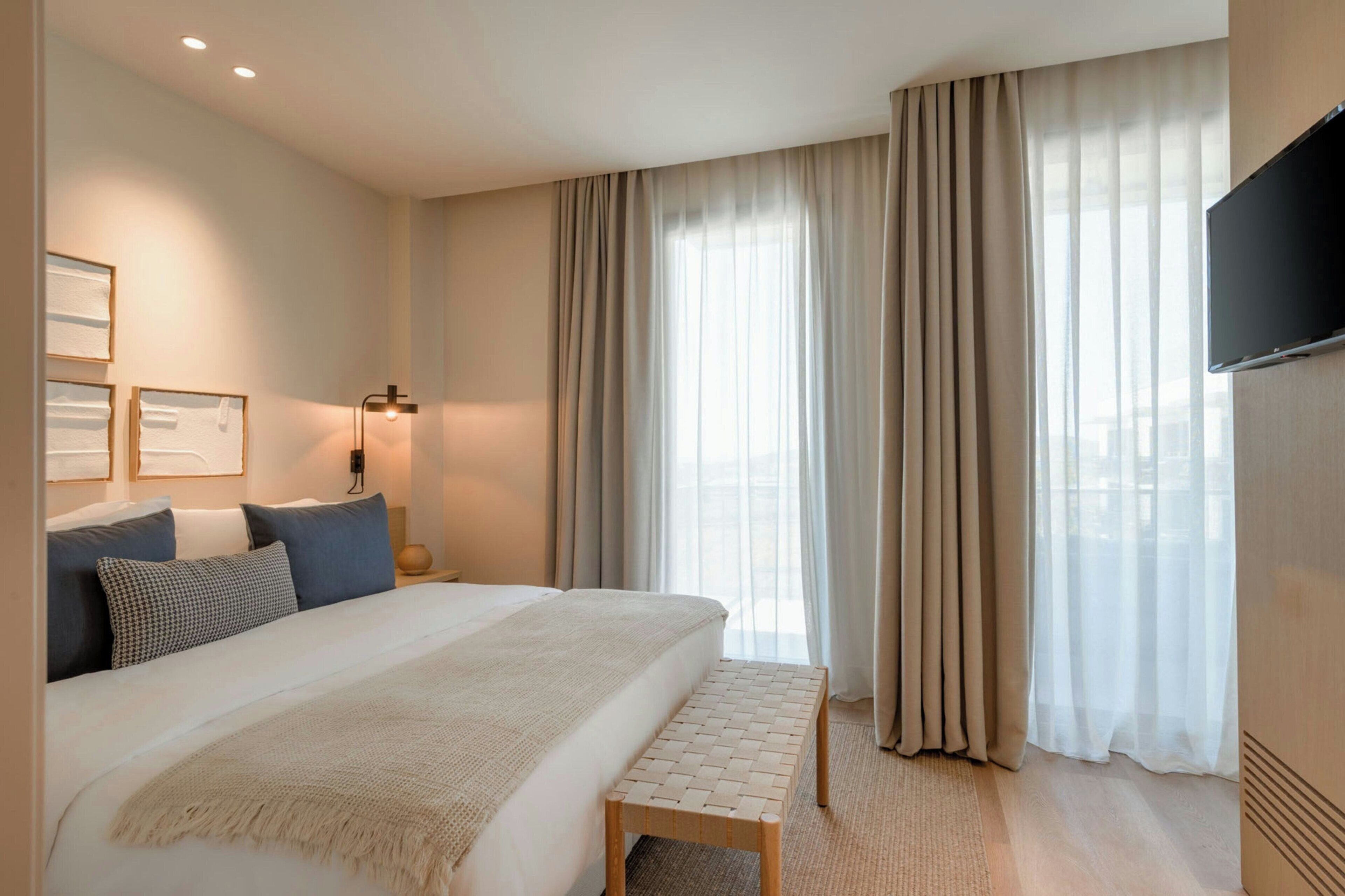 Habitación de hotel con una cama grande, una mesita de noche, un banco, un televisor y una ventana con cortinas.
