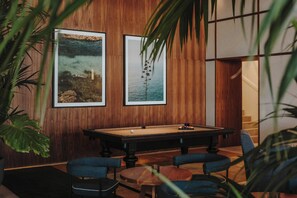 A pool table in a room with wooden paneling, two framed pictures on the wall, and a small round table with chairs.