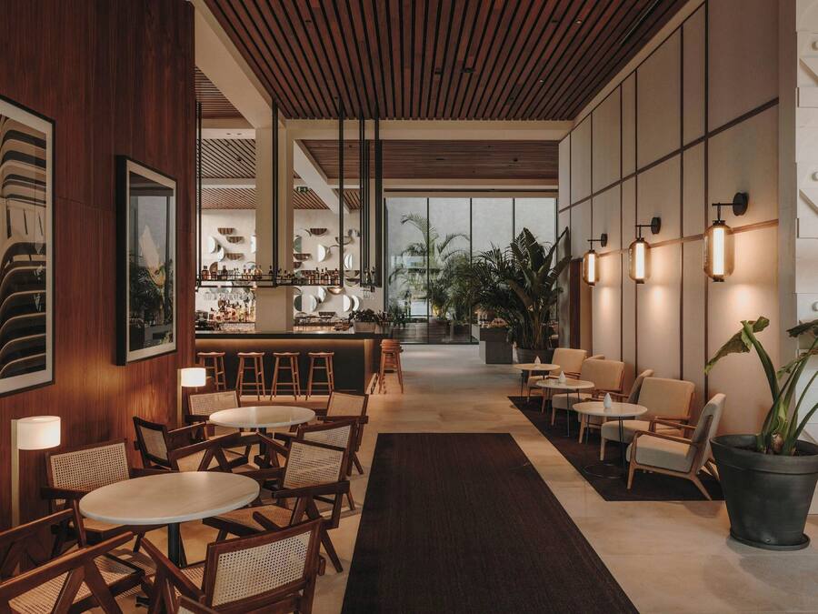 Un moderno hall de hotel con paneles de madera, mesas y sillas redondas, una zona de bar y grandes ventanales con vistas.