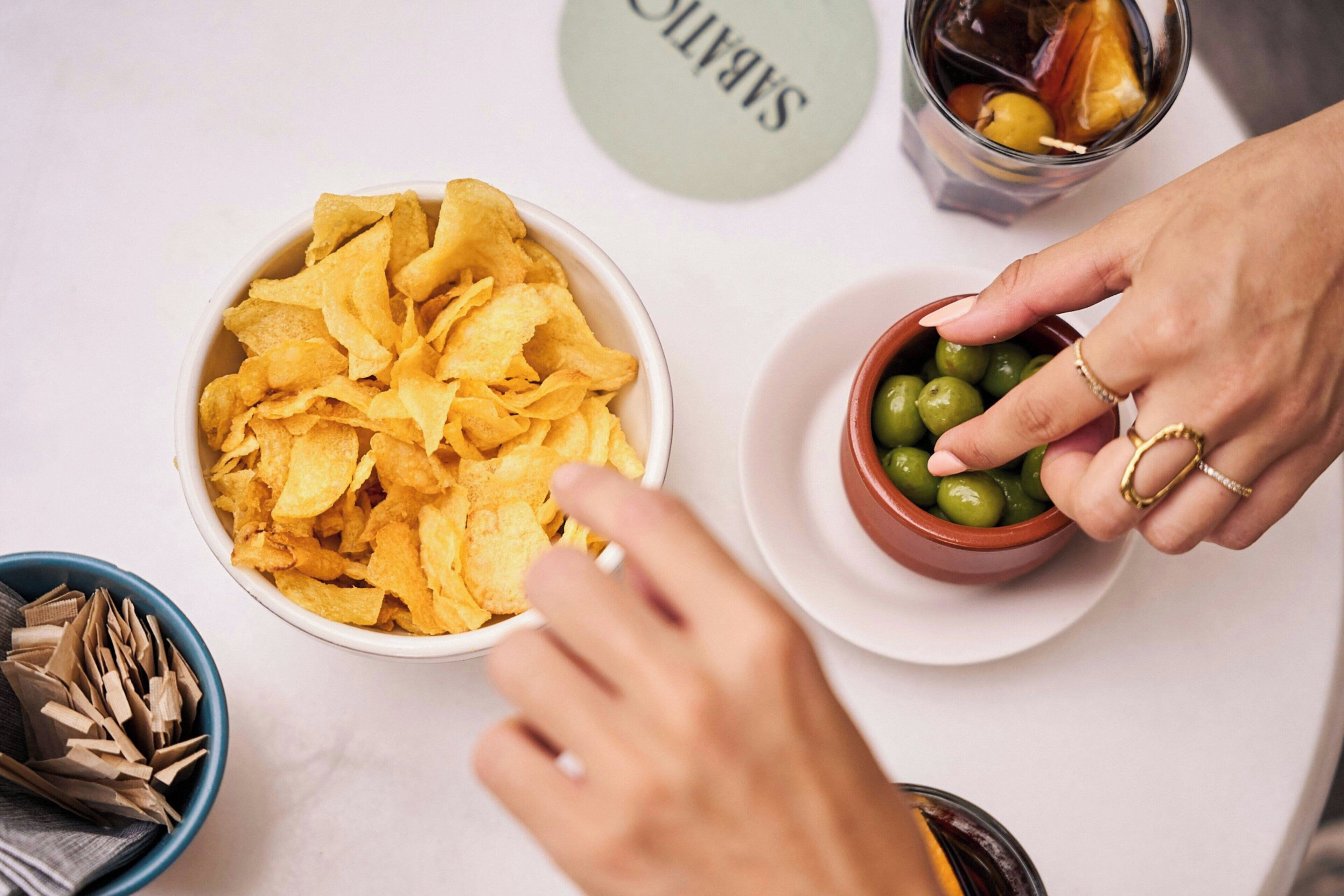 Une table avec un bol de chips, un bol d’olives et un verre de boisson fraîche.