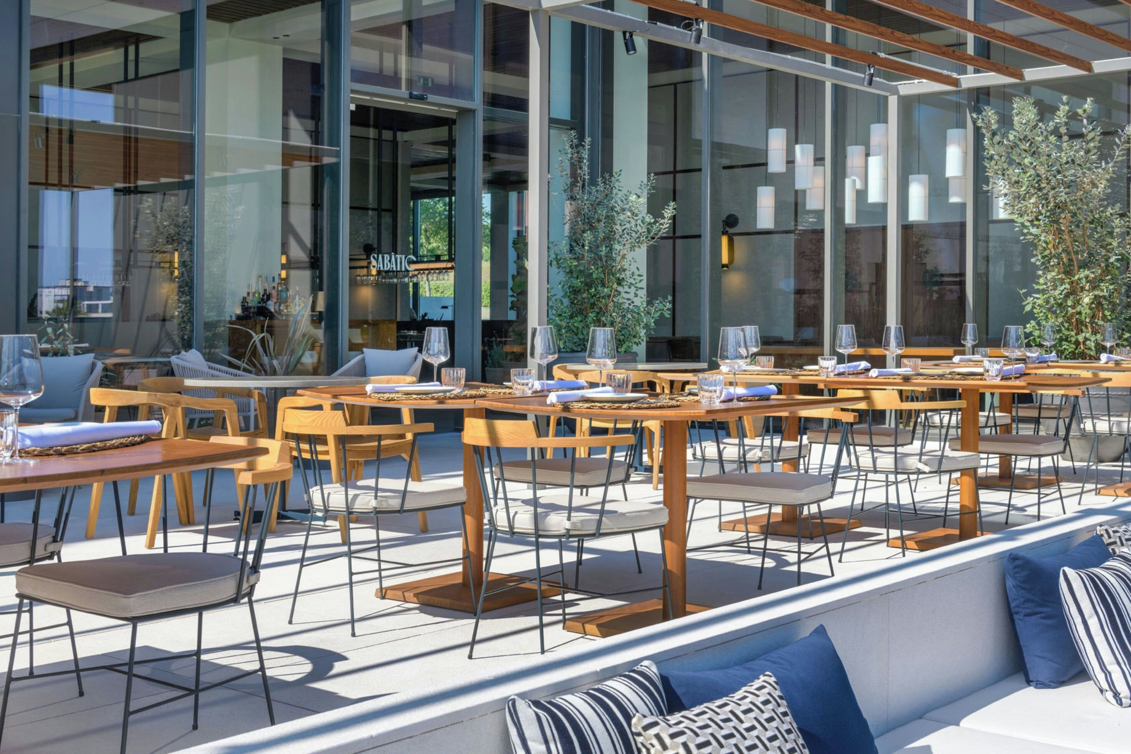 An outdoor seating area with tables and chairs, large windows, and indoor seating visible through the glass.