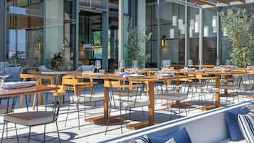 An outdoor seating area with tables and chairs, large windows, and indoor seating visible through the glass.