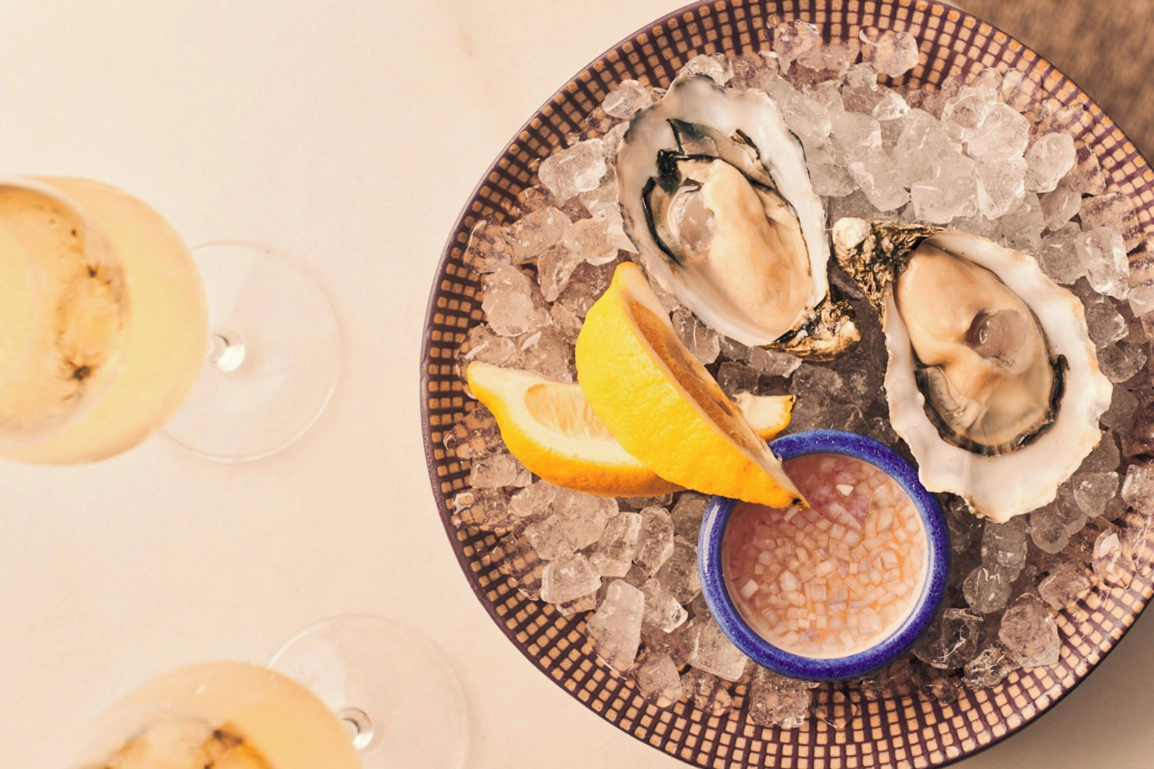 A plate of oysters on ice with lemon wedges and a blue sauce container.