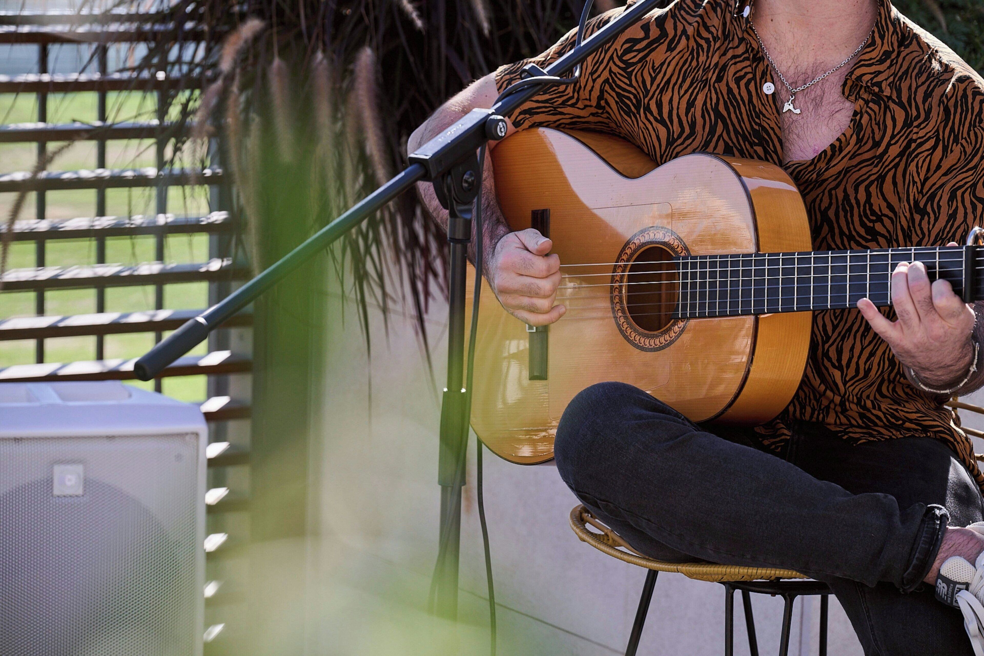 Une personne joue de la guitare acoustique assise sur une chaise, à l'extérieur.