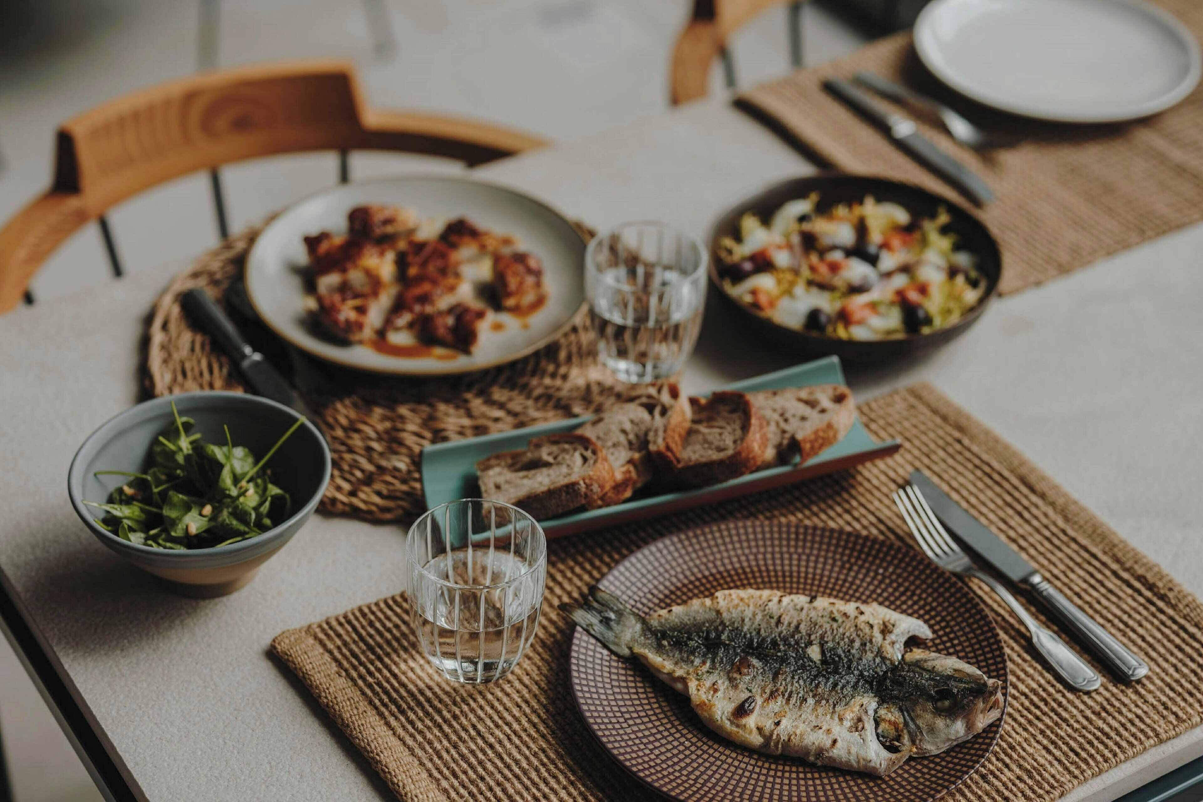 Una mesa de comedor con varios platos, entre ellos pescado, ensalada y pan.