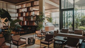 Un área de lounge moderna con disposición de asientos, una estantería de madera y grandes ventanales que dejan entrar luz natural.