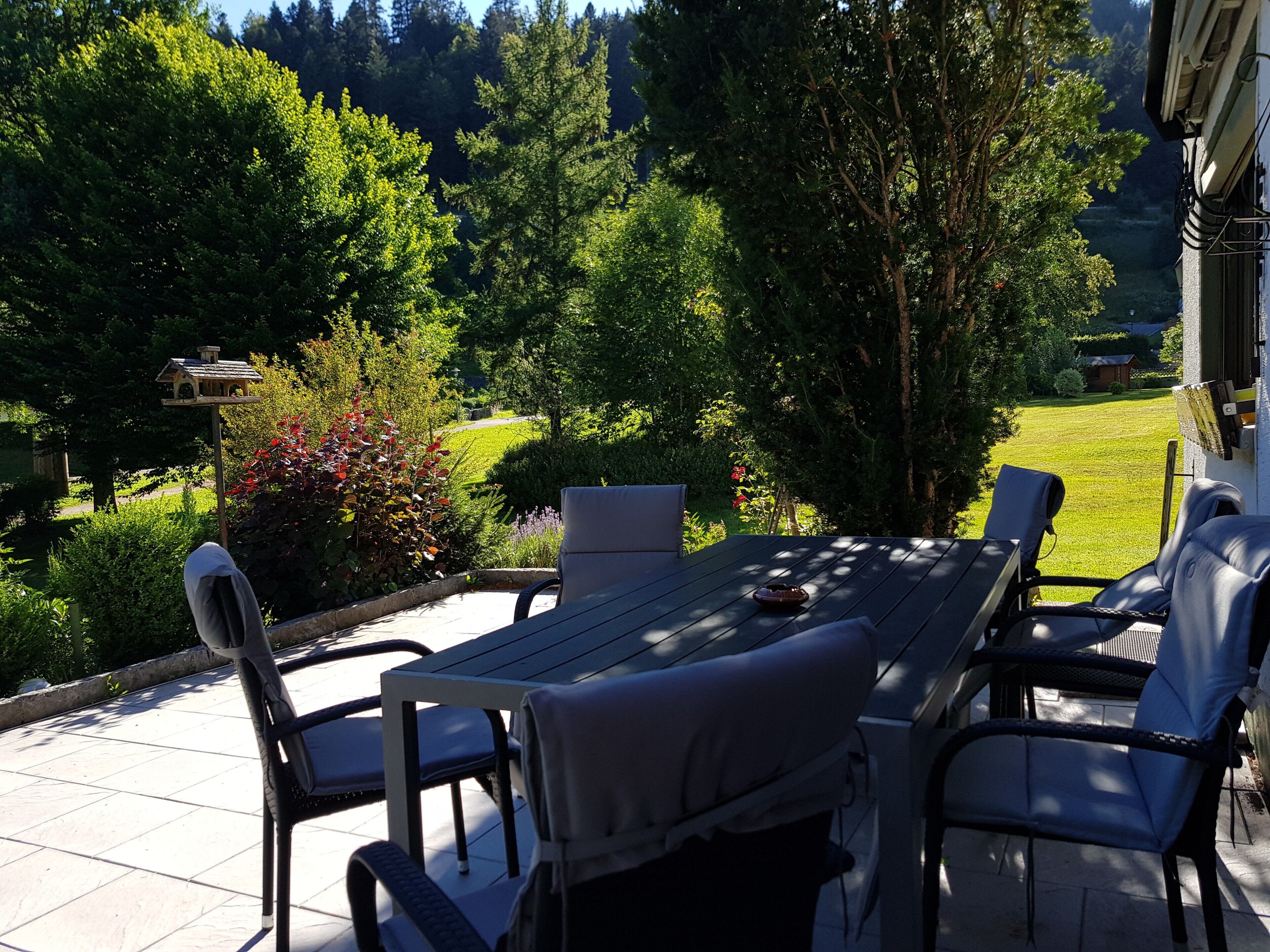 Un patio al aire libre con una mesa y sillas, rodeado de abundante vegetación y vistas a un área boscosa.