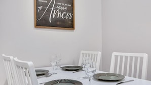 A dining table with white chairs, set with plates, glasses, and cutlery. A framed sign with the text "NOBODY EATS 'TIL WE SAY Amen" hangs on the wall. - Barnwood Suite · Romantic getaway on Main Street - Barnwood Suite (Berlin)