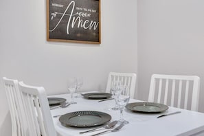 A dining table with white chairs, set with plates, glasses, and cutlery. A framed sign with the text "NOBODY EATS 'TIL WE SAY Amen" hangs on the wall.