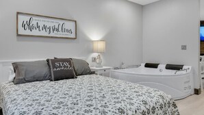 A bedroom with a large bed, a wall-mounted sign, a bedside table with a lamp, and a bathtub. - Barnwood Suite · Romantic getaway on Main Street - Barnwood Suite (Berlin)