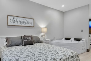 A bedroom with a large bed, a wall-mounted sign, a bedside table with a lamp, and a bathtub.
