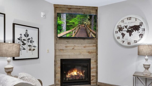A living room with a fireplace, a television mounted above it, and a world clock on the wall. - Barnwood Suite · Romantic getaway on Main Street - Barnwood Suite (Berlin)