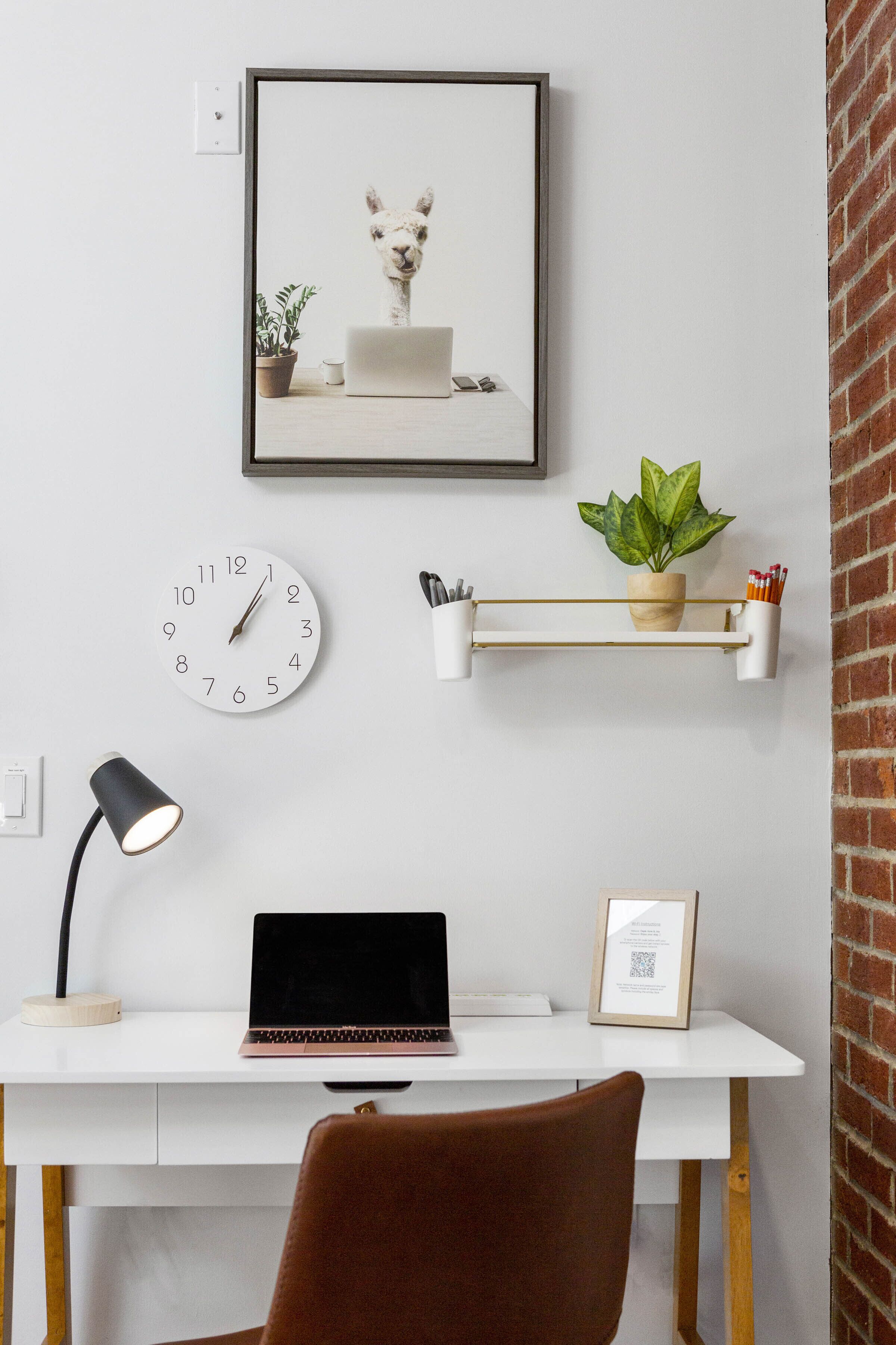 Un espacio de trabajo con una computadora portátil, un reloj, un cuadro enmarcado, una lámpara de escritorio y una planta en un estante.