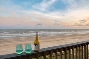 Une terrasse en bois avec une bouteille de vin et deux verres, donnant sur une plage, l’océan et un ciel au coucher du soleil.