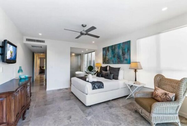 A modern bedroom with a white bed, a ceiling fan, a painting, and a wicker chair.