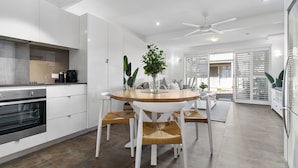 A modern kitchen with white cabinetry, a built-in oven, and a dining area with a wooden table and white chairs. - Experience the laid-back Noosa lifestyle! (Noosa)