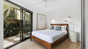 Un dormitorio con una cama de madera, un ventilador de techo, una mesita de noche con lámpara y vista a un jardín a través de una puerta corrediza de vidrio.