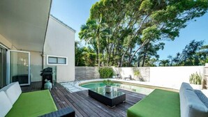 Un patio moderno al aire libre con piscina, asientos acolchados verdes y una mesa de vidrio.