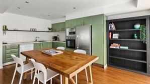 Una cocina moderna con armarios verdes, una mesa de comedor de madera, sillas blancas y una estantería negra.