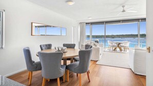 A dining area with a round table and grey chairs, a white sofa, and a view of the sea through large windows.