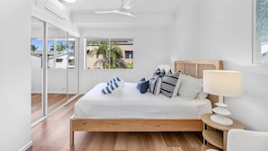 A bedroom with a bed, a ceiling fan, a bedside table with a lamp, and a view of palm trees through large windows.