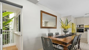 A dining area with a wooden table, four chairs, a vase with yellow flowers, and a large mirror on the wall. - Light, bright and airy with breathtaking views of Noosa Heads! (Noosa)