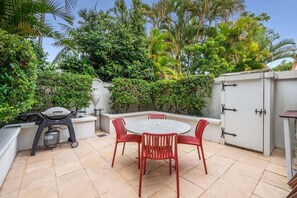 Eine Terrasse im Freien mit einem runden Tisch und roten Stühlen, umgeben von üppiger Bepflanzung und einer weißen Mauer mit einem Metallschrank.