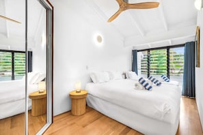 Moderni hotellihuone, jossa on suuri sänky, kattotuuletin, suuri ikkuna verhoilla sekä pieni yöpöytä lampun kanssa.