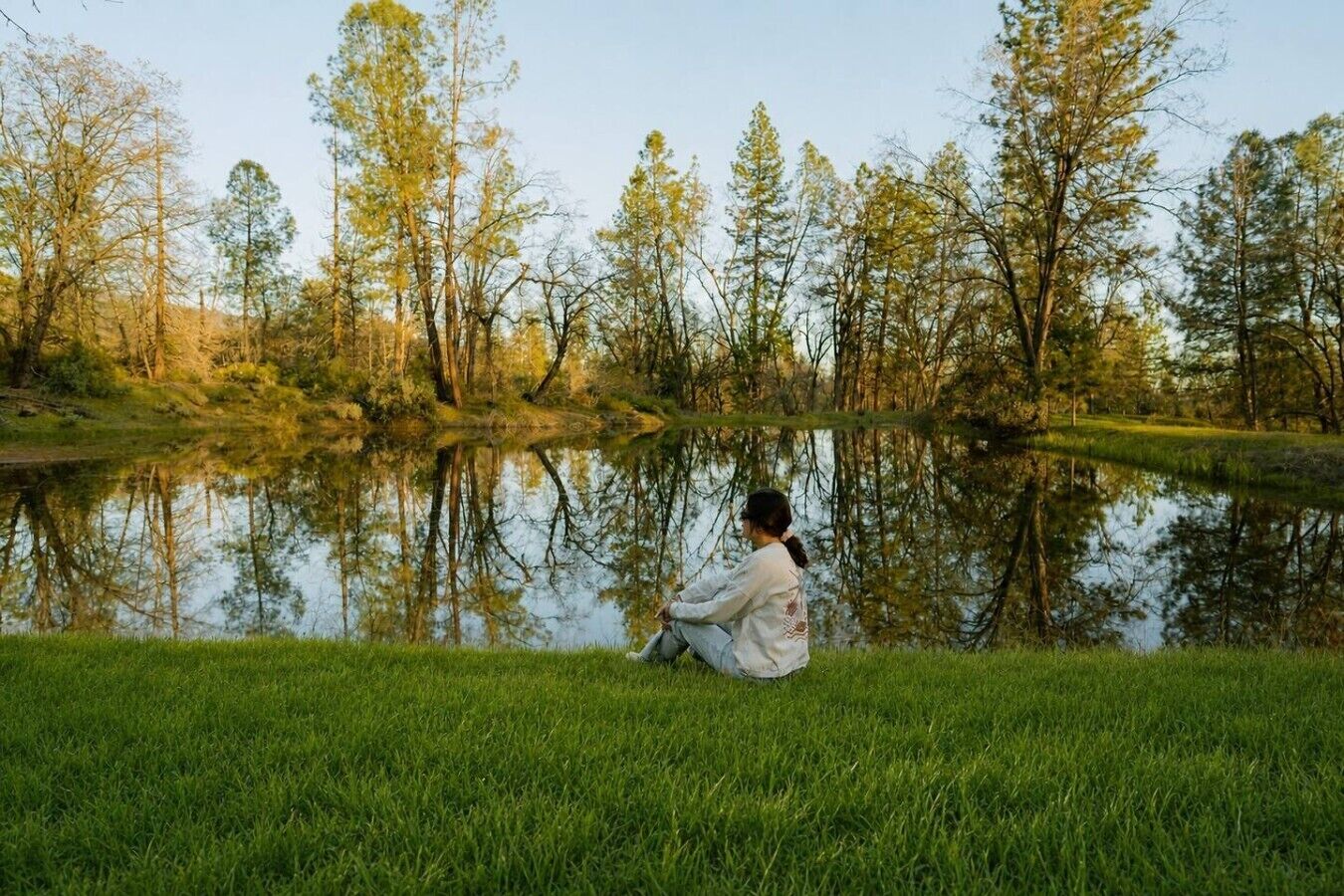 Uma pessoa sentada à beira de um lago calmo, rodeada de árvores.