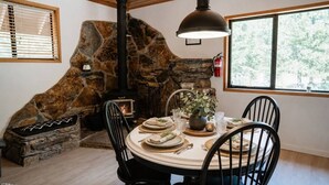 Un coin repas agrémenté d’une cheminée en pierre, d’une table ronde dressée pour quatre personnes et d’une grande fenêtre offrant une vue sur les arbres.