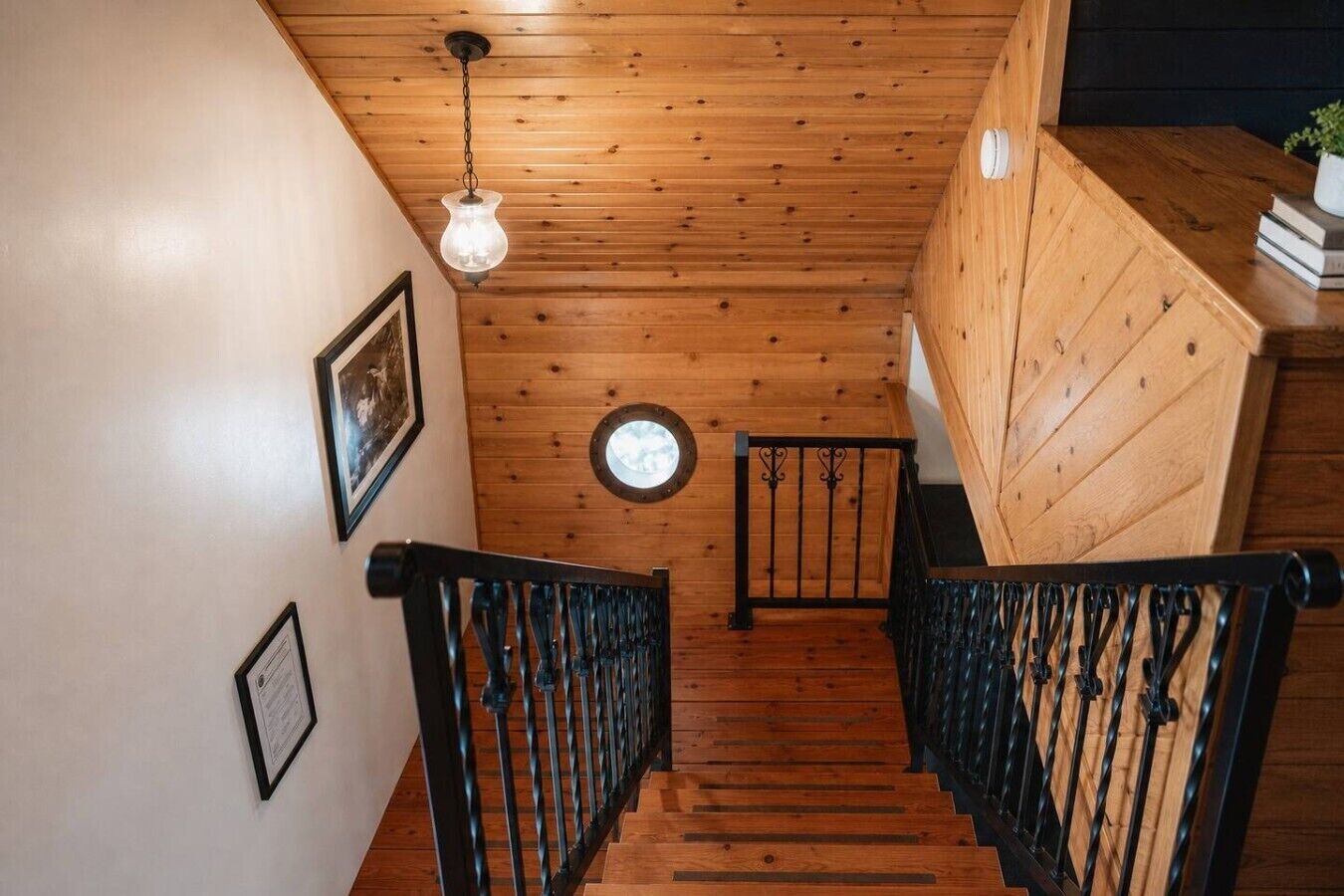 Un escalier en bois avec une rampe noire, un luminaire suspendu et une fenêtre ronde dans le mur.